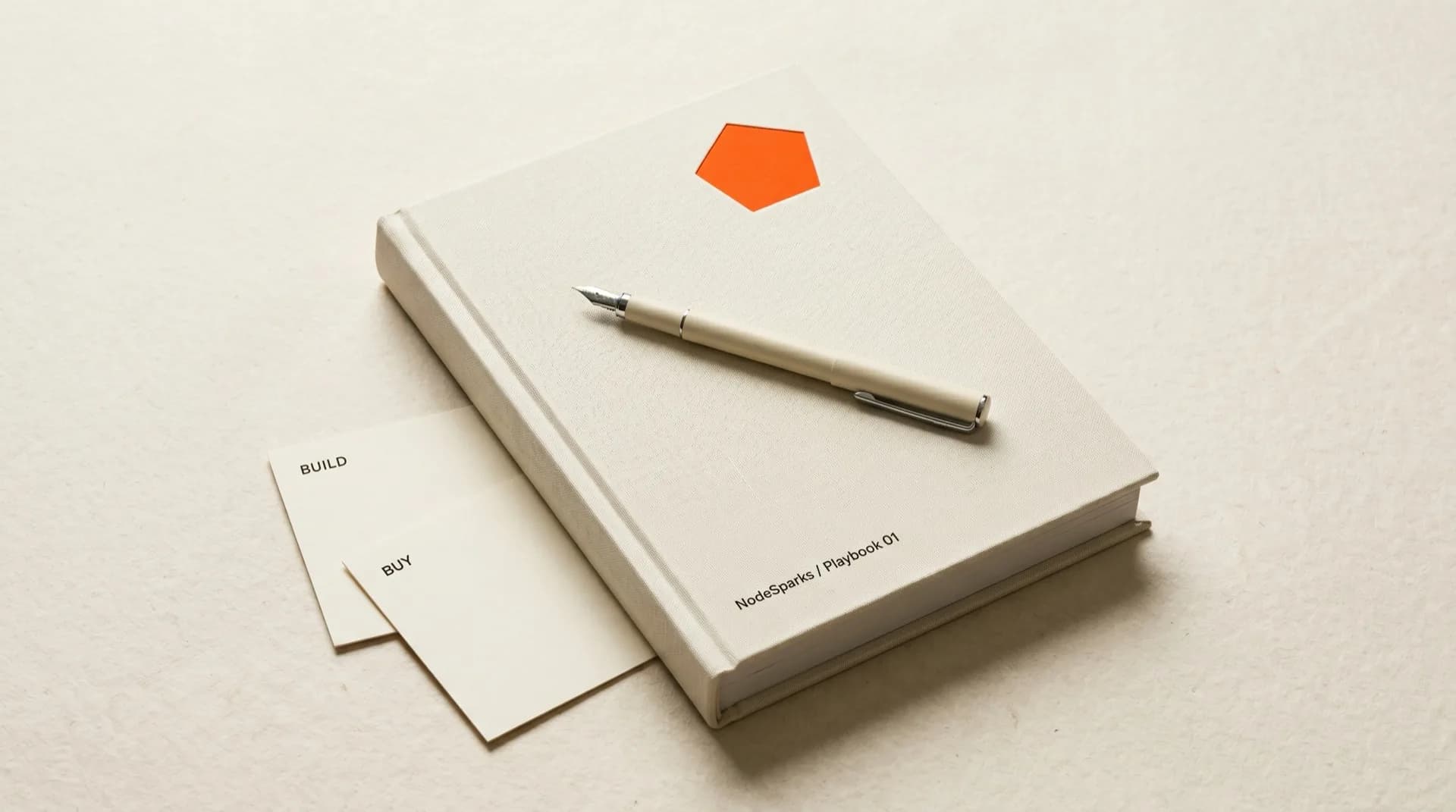 Cream linen-bound NodeSparks Playbook manual with orange pentagon emboss, build and buy index cards beside it on cream paper